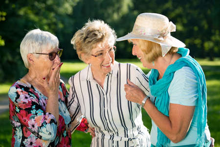 Three elderly female friends gossiping outdoors, standing in park. Shocked or surprised elderly woman holding her hand to her open mouth as she stares wide eyed at the blonde womanの写真素材