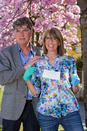 Affectionate senior couple posing holding a surgical face mask in gloved hands as protection against the Covid-19 pandemic in front of colorful pink spring blossom.の写真素材