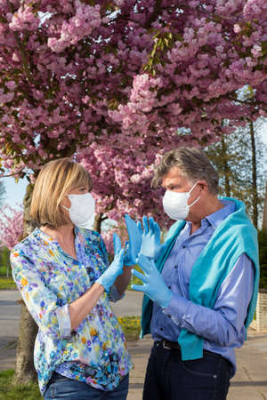 Senior couple wearing surgical face masks and gloves outdoors in spring during the coronavirus pandemic standing looking into each others eyes with love with raised hands.の写真素材