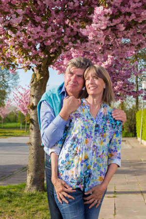 Loving elderly couple enjoying the spring sunshine standing in a close embrace with faces to the sun in front of colorful pink blossom on a tree.の写真素材