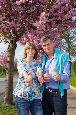 Motivated positive senior couple giving a double thumbs up with a happy smile as they stand close together in a tree lined street against pink spring blossom.の写真素材
