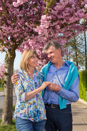 Affectionate senior couple outdoors in spring posing holding hands looking lovingly at each other under a tree covered in pink blossom.の写真素材