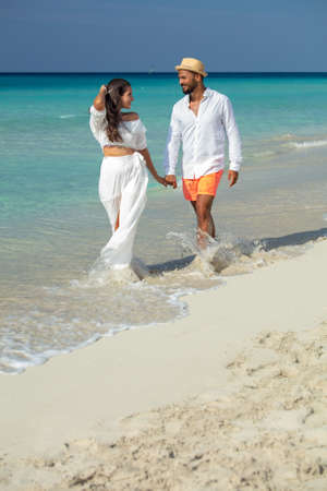 Happy romantic young couple on a honeymoon or date walking along a tropical beach at the edge of the sea, smiling into each others eyes.の写真素材