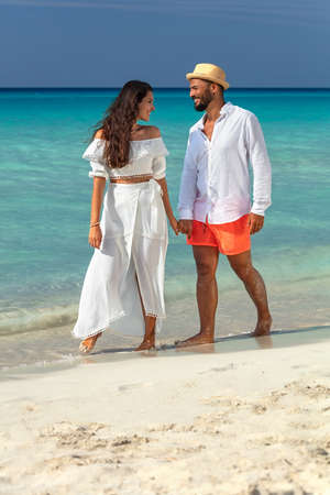 Full length of happy couple in white summer clothes walking on beach holding hands and enjoying their honeymoon. Man and woman looking at each other and smiling.の写真素材