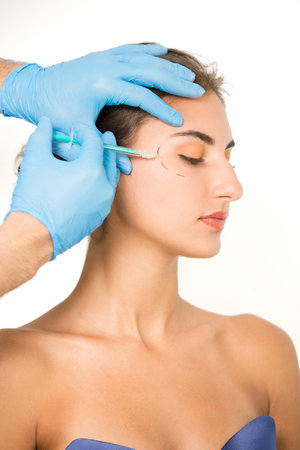 Hands of male cosmetician inserting syringe on markings over young woman's face against white background.の写真素材