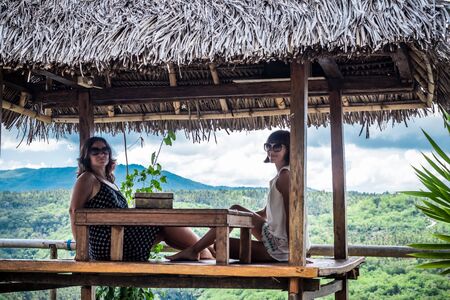 Two Beautiful happy young girls sitting in a wooden gazebo at sunny day. and having fun, smiling and laughing. Tropical Bali island.の写真素材