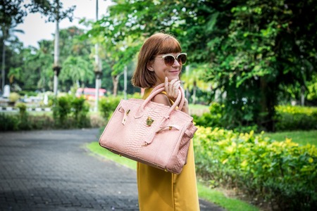 Portrait of a Beautiful fashionable caucasian brunette woman with sunglasses and luxury snakeskin python handbag posing in the park of Nusa Dua, Bali, Indonesia. Asia. Sunny day, green background.の写真素材