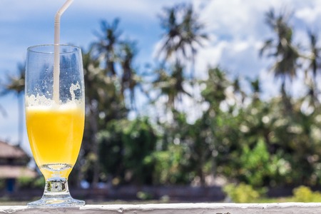 Glass of orange juice on bright tropical blurred background with palms, outdoors. Tropical Bali island, Indonesia.の写真素材