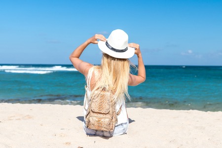 Young sexy woman with white hat and luxury snakeskin python backpack sitting on white sand beach a tropical Bali island at sunny day. Ocean cost.の写真素材