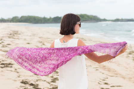 Portrait of young sexy attractive woman in white dress with silk scarf alone on the tropical beach of Bali Island, Indonesia.の写真素材