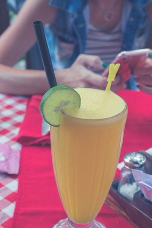 Glass of fresh orange juice with slice of lime on red background in restaurant. Tropical Bali island, Indonesia. Young woman with smartphone on a blurred background.の写真素材