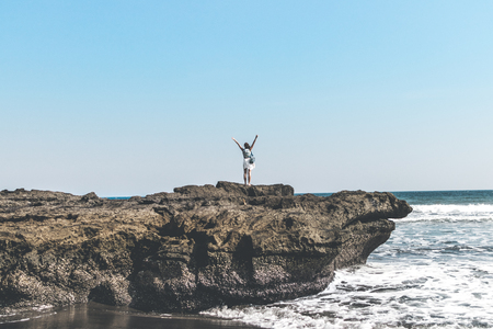 Young woman on the rock on a black sand beach, Bali island. Freedom concept.の写真素材