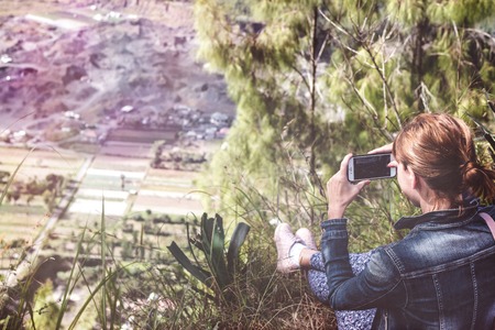 Successful woman hiker taking picture with smartphone at cliff edge on mountain top. Bali island.の写真素材