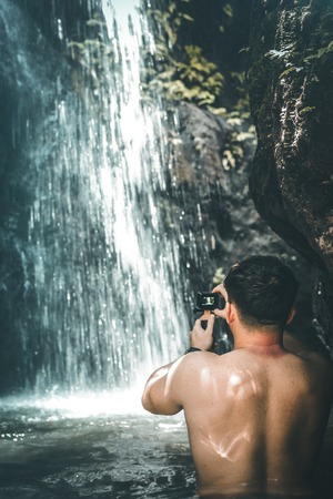 Tourist man on a waterfall background holds an action camera and takes a picture. Bali island.の写真素材