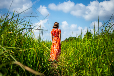 Young woman in red dress walking on a trail with long green grass. Bali island. Indonesia. Asia.の写真素材