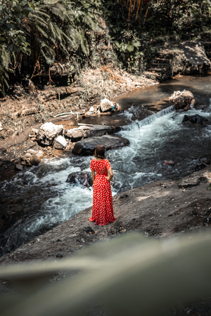 Young woman in long red dress posing on a mountain river deep in the rainforest of Bali island.の写真素材