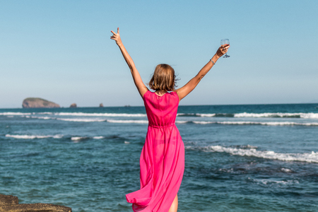 Young woman in a long red dress and with a glass of wine posing on the sea background.の写真素材