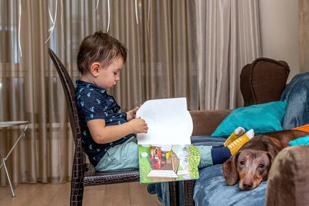 MOSCOW, RUSSIA - OCTOBER 12, 2019: Little 3 years boy and dachshund dog reading book at home.のeditorial素材