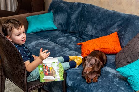 MOSCOW, RUSSIA - OCTOBER 12, 2019: Little 3 years boy and dachshund dog reading book at home.のeditorial素材