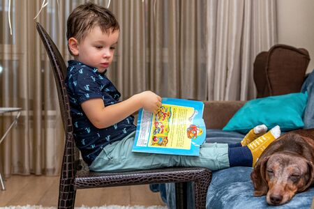 MOSCOW, RUSSIA - OCTOBER 12, 2019: Little 3 years boy and dachshund dog reading book at home.のeditorial素材