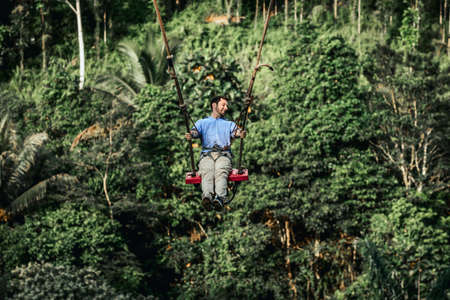BALI, INDONESIA - MAY 16, 2018: Young man swinging in the jungle rainforest of Bali island, Indonesia. Swing in the tropics. Swings - trend of Bali island.のeditorial素材