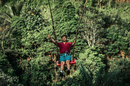 BALI, INDONESIA - MAY 16, 2018: Young man swinging in the jungle rainforest of Bali island, Indonesia. Swing in the tropics. Swings - trend of Bali island.のeditorial素材