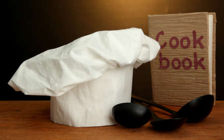 Chef's hat with spoons and cookbook on table on brown backgroundの写真素材