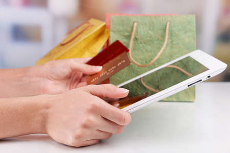 Female hands holding credit card with computer tablet and paper bags on table on bright backgroundの写真素材