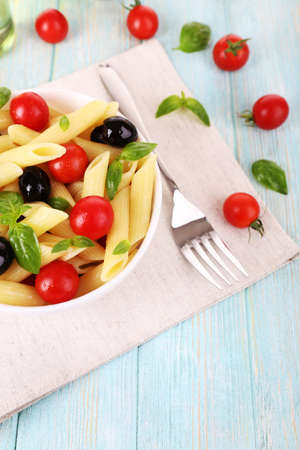 Pasta with tomatoes, olives and basil leaves in bowl on napkin on wooden backgroundの写真素材