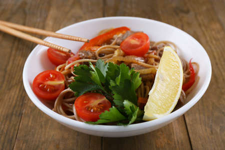 Chinese noodles with vegetables and roasted meat in bowl on napkin, on wooden backgroundの写真素材