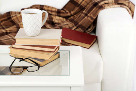 Tabletop with pile of books, cup and glasses near the sofa with plaidの写真素材