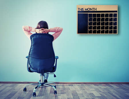 Woman sitting back on the office chair and looking at chalkboard calendar on wall backgroundの写真素材