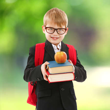 Little boy with backpack holding books and apple on blurred green background. School concept.の写真素材