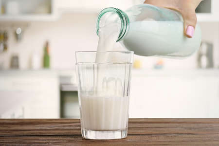Female hand pouring milk into glass against blurred kitchen background. Dairy concept.の写真素材