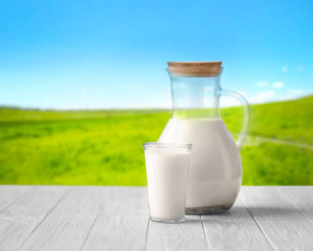 Glass and jug with milk on white wooden table against blurred nature background. Dairy concept.の写真素材