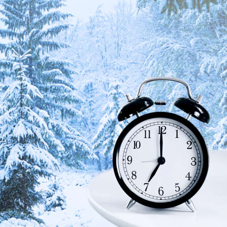 Alarm clock on table against beautiful snowy landscape. Daylight saving time concept.の写真素材
