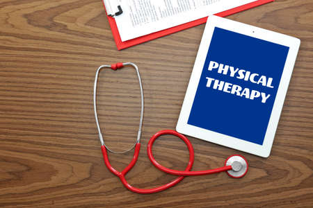 Rehabilitation concept. Tablet and stethoscope on wooden background. Text PHYSICAL THERAPY on screenの写真素材
