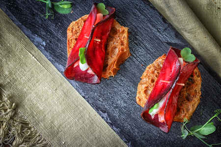 Toasts with a circle of beets, goat cheese and a piece of salmon on a black slate on a background of rustic napkin.Decorated with shoots of green peas leaves.の写真素材