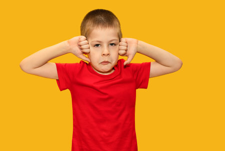 Child in red t-shirt on yellow background, boy 5 years old, sad. Shows fingers down. Sad children's emotionsの写真素材