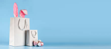 Wide banner on a blue background. The concept of online shopping for Easter, purchases and sale for the holiday. Gift bag with bunny ears and Easter eggsの写真素材