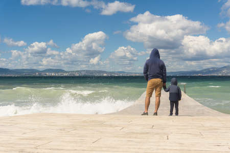 Father and son are standing on the pier, admiring the seascape, raging autumn sea. Man holding a child by the handの写真素材