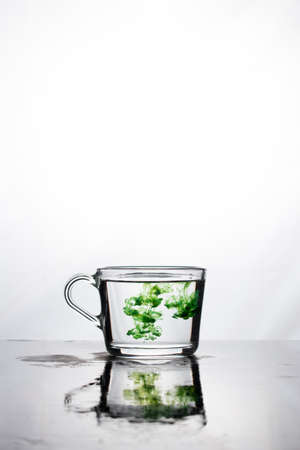 Green color dissolves in a cup of water on a white background.The mug is reflected on the wet surface. At the top there is space for text. vertical photo.の写真素材