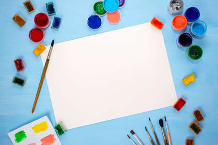 Watercolor, acrylic, brushes and a white sheet of paper on a blue background. Flat lay, top view. Minimalistic desktop of the artist. Preparation for drawing.の写真素材