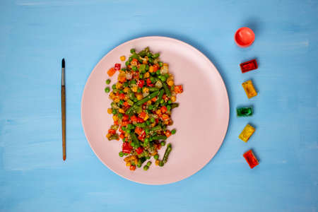 red, green and yellow fried vegetables on a pink plate. on the sides of the plate are an art brush and palettes of colorful paints. blue background. concept of drawn foodの写真素材