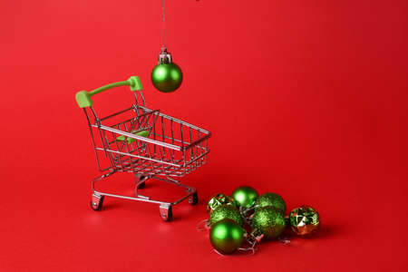 Female hand put green Christmas-tree decorations in a small supermarket trolley on a red background.New Year concept. Horizontal photo. Place for text.の写真素材