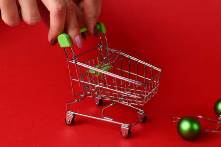 An empty small trolley drives up to a green Christmas tree toy on a red background. Female hands hold the trolley. New Year shopping concept on a red background. close-upの写真素材