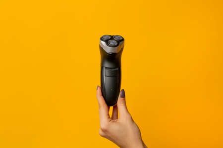 female hand holds a male electric razor on a yellow background. Minimal style.の写真素材