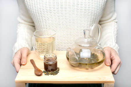 a girl in a white sweater holds a wooden tray with a glass teapot, a mug, a spoon and a jar of honey. Oolong tea is brewed in the teapot.の写真素材