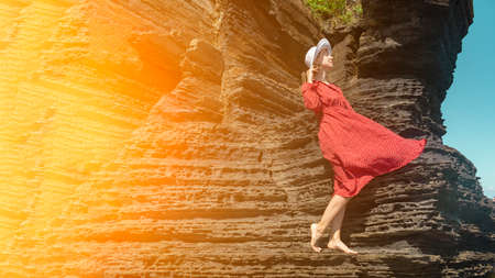 A woman in a red dress and white hat stands on a rock and looks at the sun. Dress flutters in the wind. The girl is resting alone. Retro photography. Summer lifestyle.の写真素材