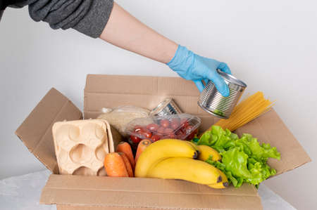 Box with food for delivery. A female hand in a medical glove puts canned food in a box with products. Concept of donation, food deliveryの写真素材